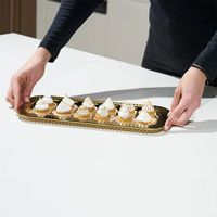 Hands carrying the Anna New York Striations Gold Small Rectangle Serving Tray with a dozen mini lemon meringue tarts on a white surface.