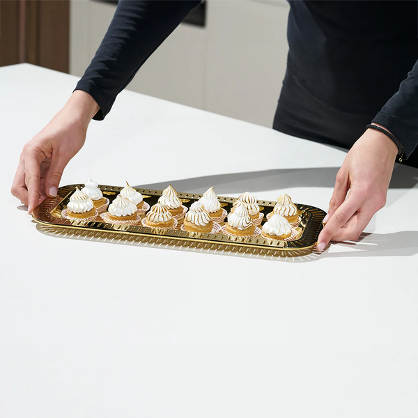 Hands carrying the Anna New York Striations Gold Small Rectangle Serving Tray with a dozen mini lemon meringue tarts on a white surface.