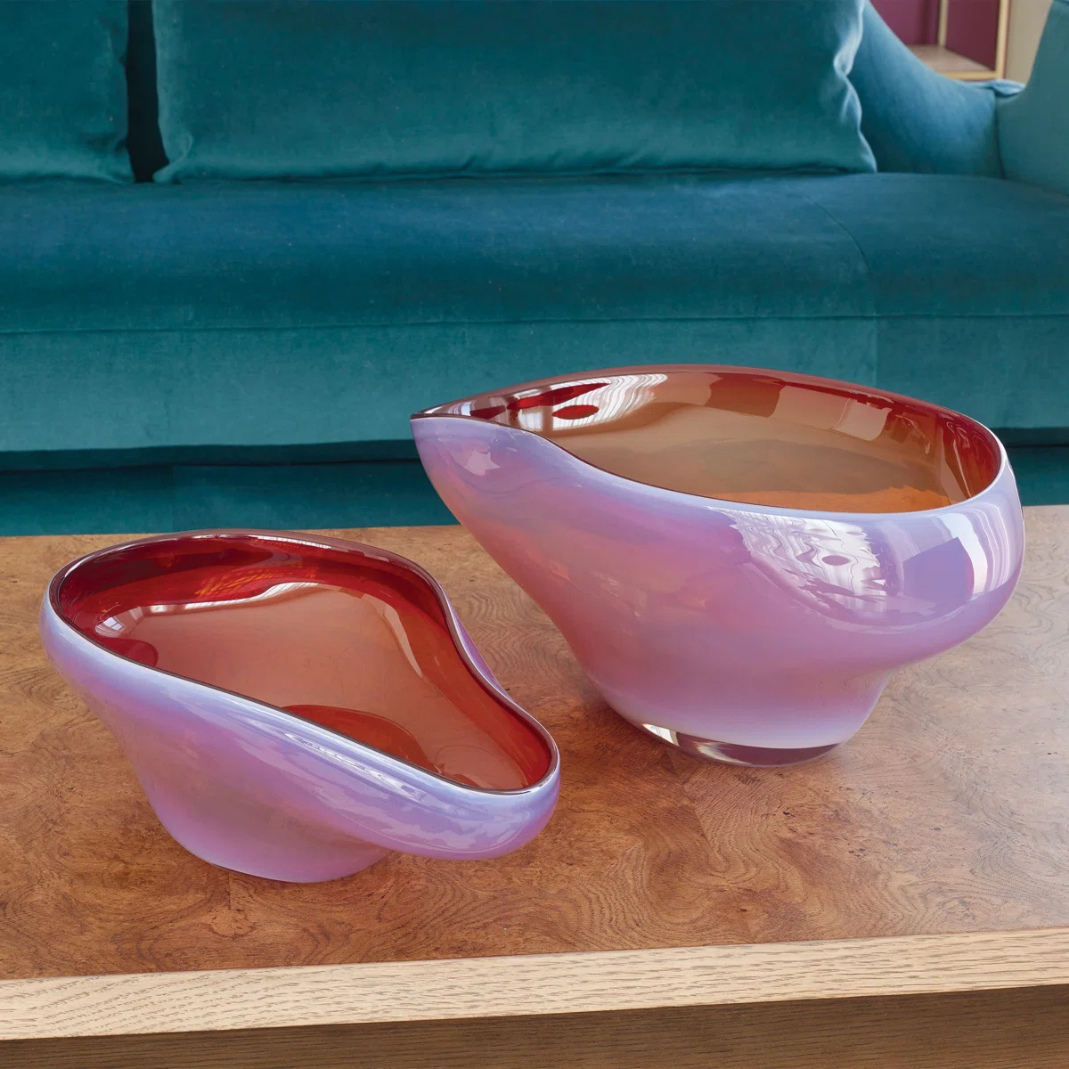 The Harmony Bowl Red - Large by Global Views/Surya, with its modern asymmetrical handblown glass design and glossy lavender-red finish, makes a striking decorative catchall on a wooden coffee table before a blue velvet sofa.