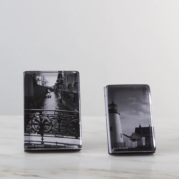 Two Simon Pearce Woodbury Photo Block Verticals sit on a marble surface. The left holds a black-and-white canal scene with a bridge, while the right displays a lighthouse and house, both in dramatic monochrome.