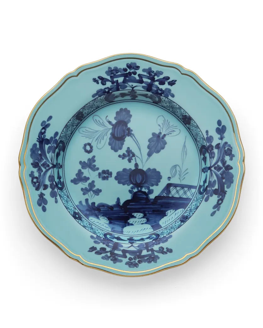 The Ginori 1735 Oriente Italiano Dinnerware Iris is a decorative light blue porcelain plate with a scalloped edge, gold trim, and intricate dark blue chinoiserie floral motifs, inspired by traditional Ginori craftsmanship.