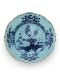 The Ginori 1735 Oriente Italiano Dinnerware Iris is a decorative light blue porcelain plate with a scalloped edge, gold trim, and intricate dark blue chinoiserie floral motifs, inspired by traditional Ginori craftsmanship.