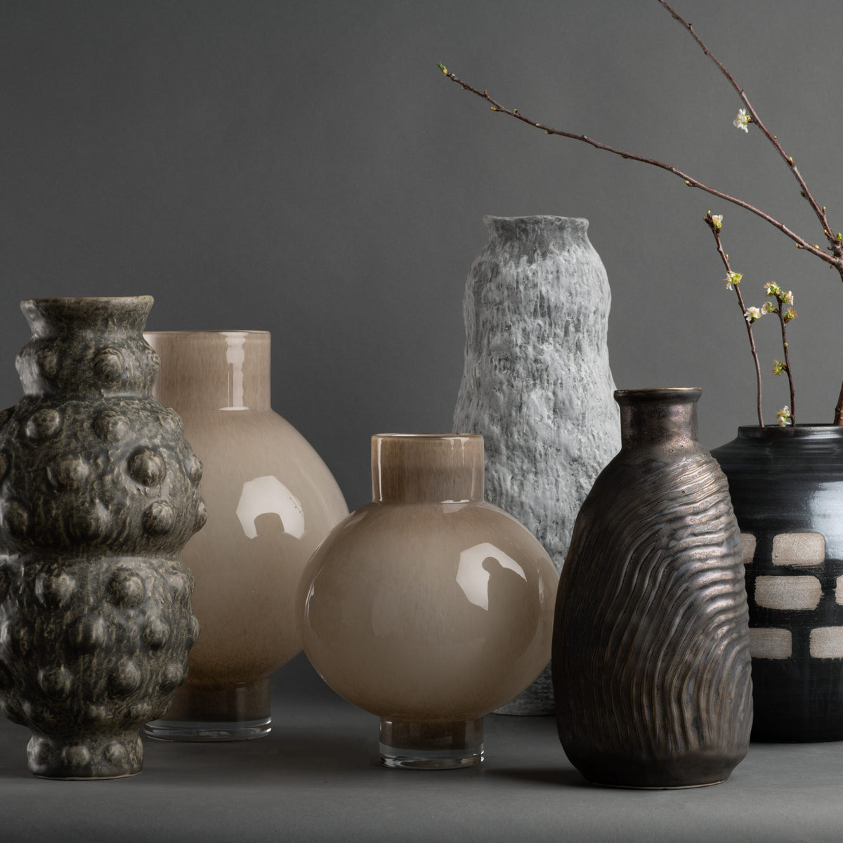 Six ceramic vases, including FlowDecor Inc.'s Tobias Vase and a contemporary design, sit on a gray surface. Their smooth, glossy, and ribbed textures add visual appeal. One vase holds thin branches with buds against a gray background.