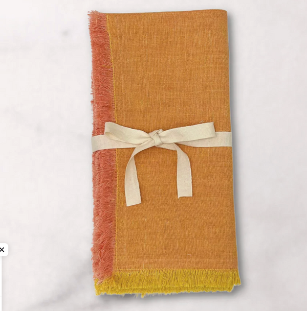 The Lia Napkin Set of 4 - Pomelo by Be Home features folded orange yellow napkins with frayed edges, pink and yellow fringe, and a cream ribbon bow, adding charm to your table setting against a light background.