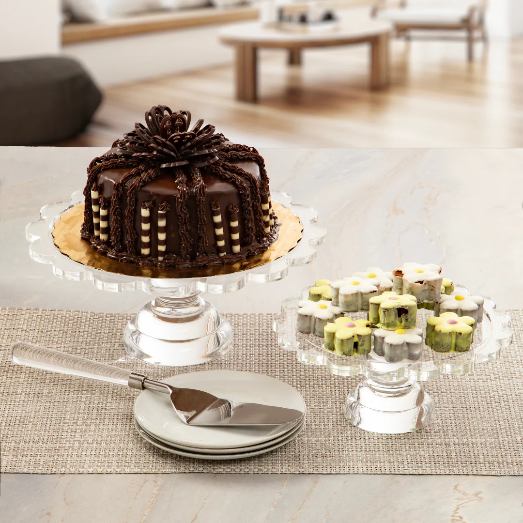 A chocolate cake adorned with decorative icing and mini pastries topped with flowers are displayed on glass stands—a clear pedestal server and a resin acrylic cake stand with scalloped edges—set beside plates and the Coco Scalloped Cake Server by Inspired Generations, all arranged on a table in a modern living room.