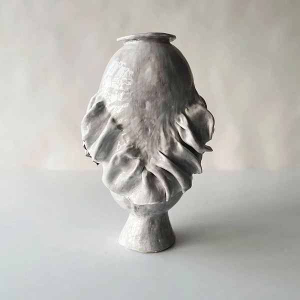 The Without Her Vase by Ellen Robinson Studio features large, wavy folds on an irregular surface with a glossy light gray finish atop a small base, highlighting its unique artistry against a neutral background.
