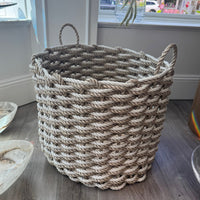 The Rope Co's Coastal Outdoor Basket in Sand & Oyster, a large round handwoven piece with thick light rope and two handles, sits on a wood floor near a window, bathed in natural light.