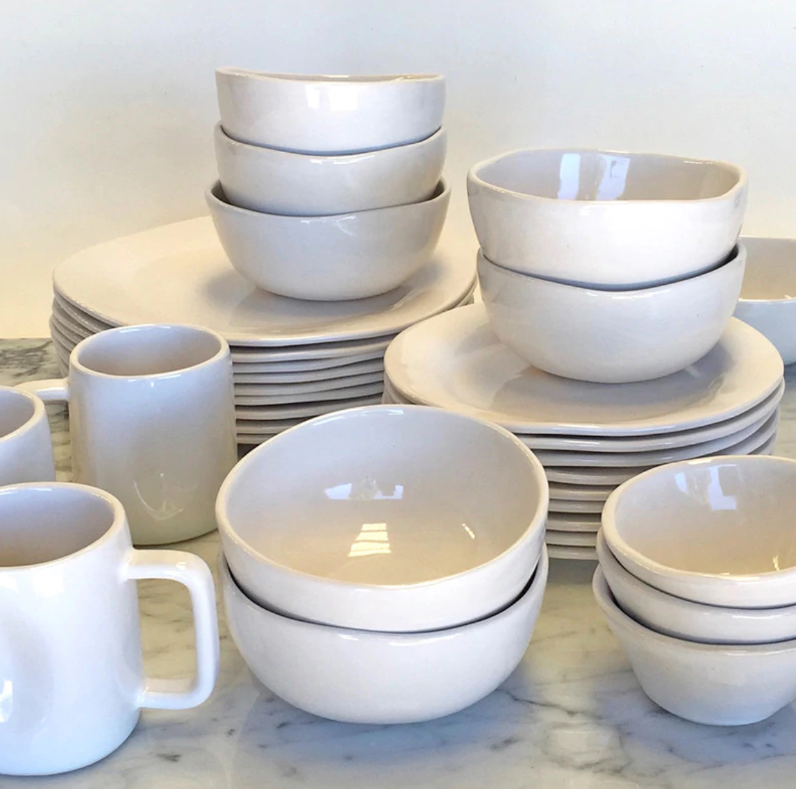 The Urban Dinnerware Matte White set by Alex Marshall Studios features handmade ceramic plates, bowls, and mugs, neatly stacked on a marble countertop against a plain background.