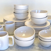 The Urban Dinnerware Matte White set by Alex Marshall Studios features handmade ceramic plates, bowls, and mugs, neatly stacked on a marble countertop against a plain background.