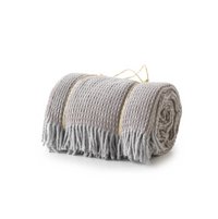 A Soft Idea’s Uniwaffle Throw, light gray with a waffle texture and fringe, is rolled up and tied with beige string, shown on a white background.
