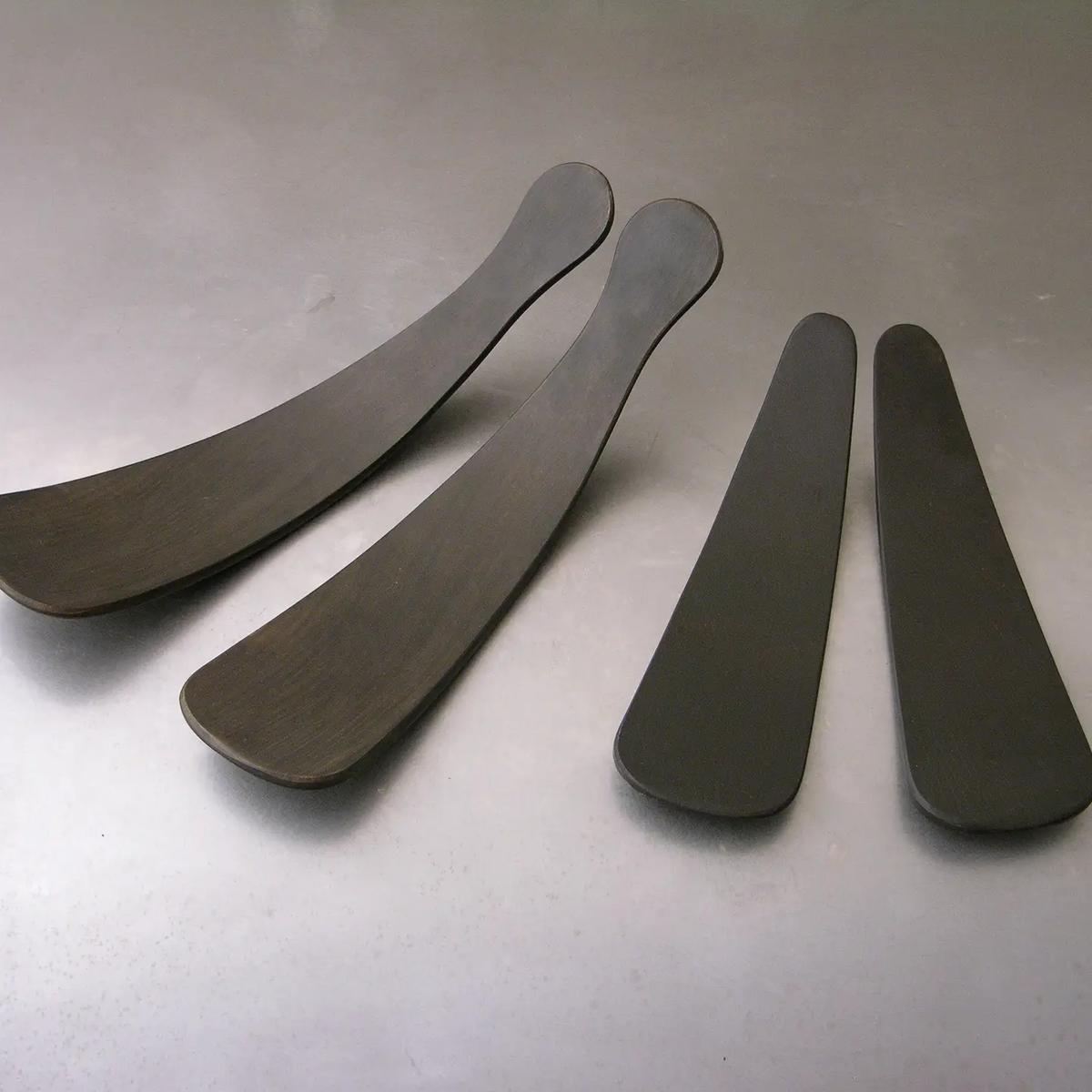 The Peterman Ebonized Oak Salad Tosser Set features four dark, curved tossers of varying lengths, with the two on the left being longer and wider than the shorter, narrower pair on the right—an elegant addition to any dining table.
