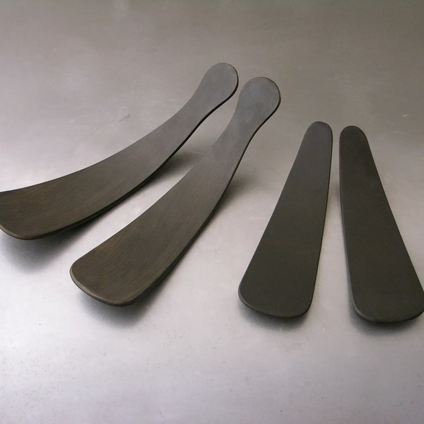 The Peterman Ebonized Oak Salad Tosser Set features four dark, curved tossers of varying lengths, with the two on the left being longer and wider than the shorter, narrower pair on the right—an elegant addition to any dining table.