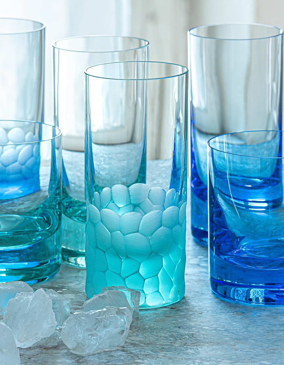 Six blue and turquoise Moser Pebbles Highball 400ml glasses, including a hand-blown glass with a pebble pattern, are arranged on a light surface with ice cubes. Light passes through the glasses, creating a fresh, cool look.
