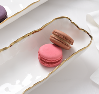 Two macarons, one pink and one brown, sit on the PAMPA BAY Portofino Small Tray with a wavy gold rim. The tray rests on a white surface beside a folded white napkin.