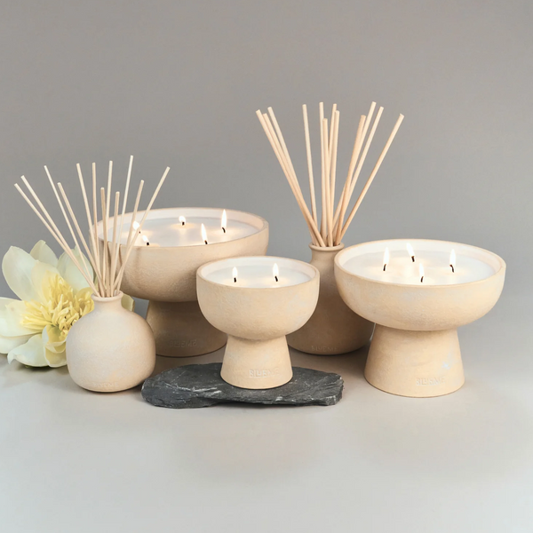 Blueme Balance Candles made from a soothing soy wax blend are arranged with reed diffusers on a gray surface, accented by a yellow flower and dark stone, set against a light gray background.