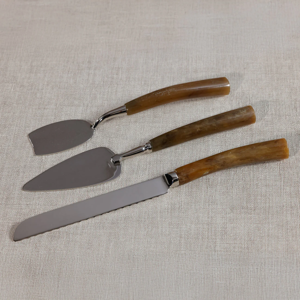 The Zodax Borghese Resin Handle 3 Piece Cheese Set—featuring a cake server, pie server, and serrated knife—rests on a light beige fabric. This elegant set makes a thoughtful host or hostess gift.