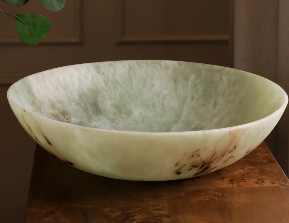 The Resin Giada Centerpiece Bowl - Jade Swirl by Beatriz Ball, featuring a shallow, round shape and marbled green design with brownish streaks, sits on a wooden surface. Green leaves are visible in the upper left corner.