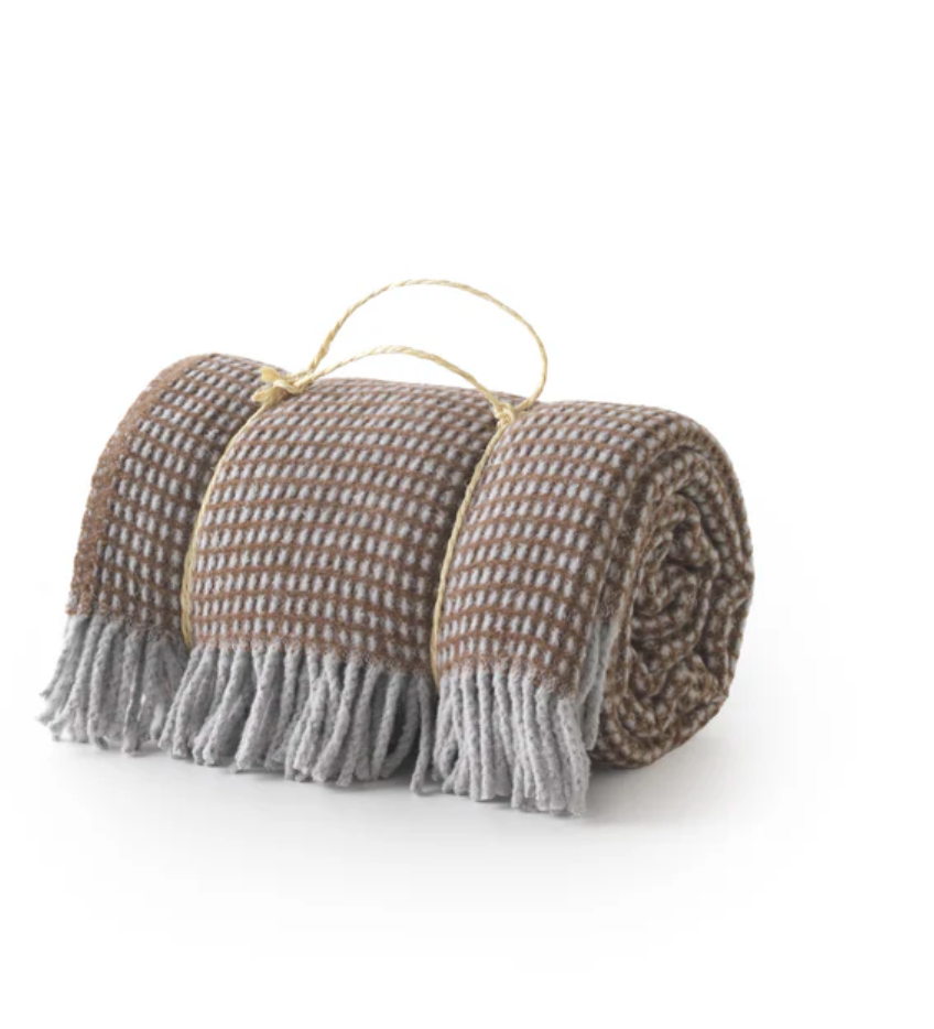 A Soft Idea's Uniwaffle Throw, a brown and gray waffle-textured blanket with fringed edges and a string handle, is shown rolled up on a white background.