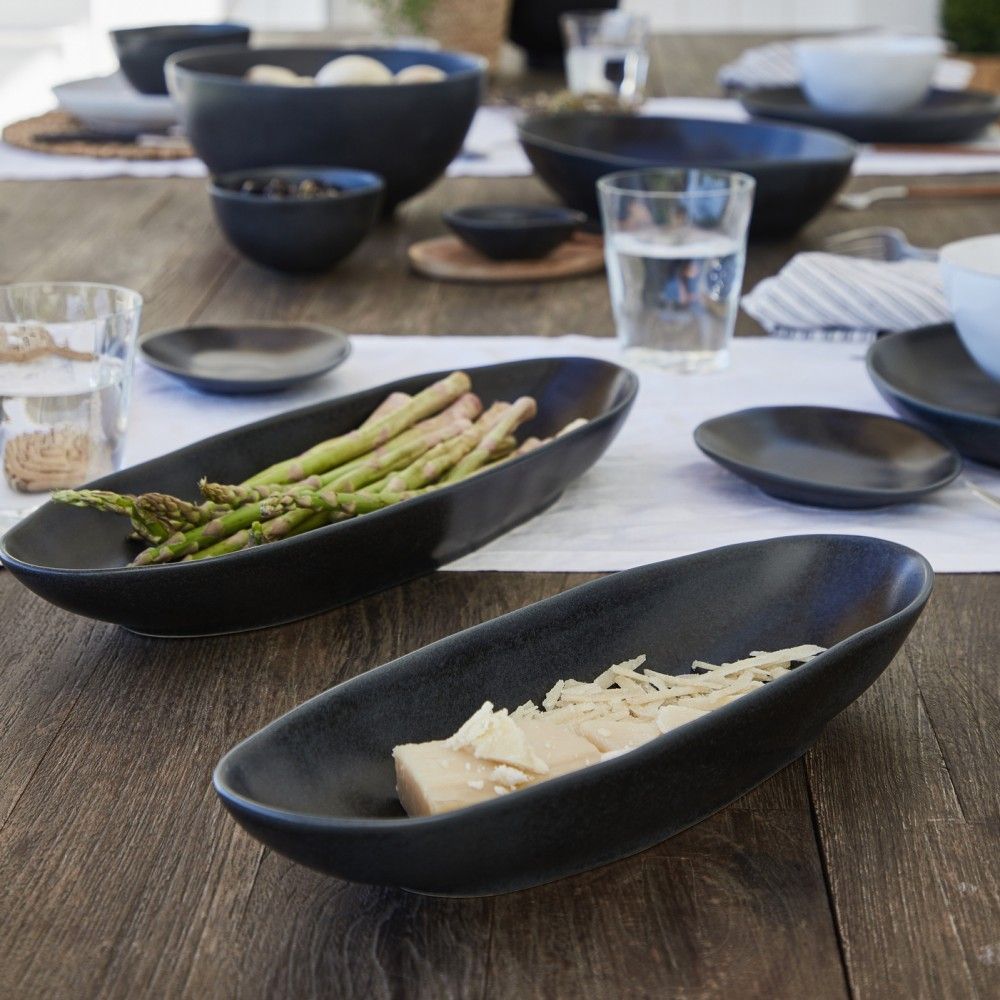 Two Livia Dinnerware Matte Black oval dishes by Casafina/Costa Nova sit on a wooden table—one with cooked asparagus, the other with cheese topped with shredded cheese. Other stoneware set pieces and glasses of water complete the scene.