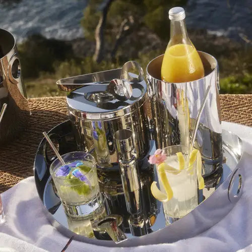 A reflective silver tray displays two garnished cocktails, an Oh De Stainless Steel Wine Cooler by Christofle holding orange juice, an ice bucket with tongs, and a jigger—ideal for a chic outdoor home bar with greenery and water in the background.