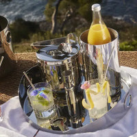 A reflective silver tray displays two garnished cocktails, an Oh De Stainless Steel Wine Cooler by Christofle holding orange juice, an ice bucket with tongs, and a jigger—ideal for a chic outdoor home bar with greenery and water in the background.