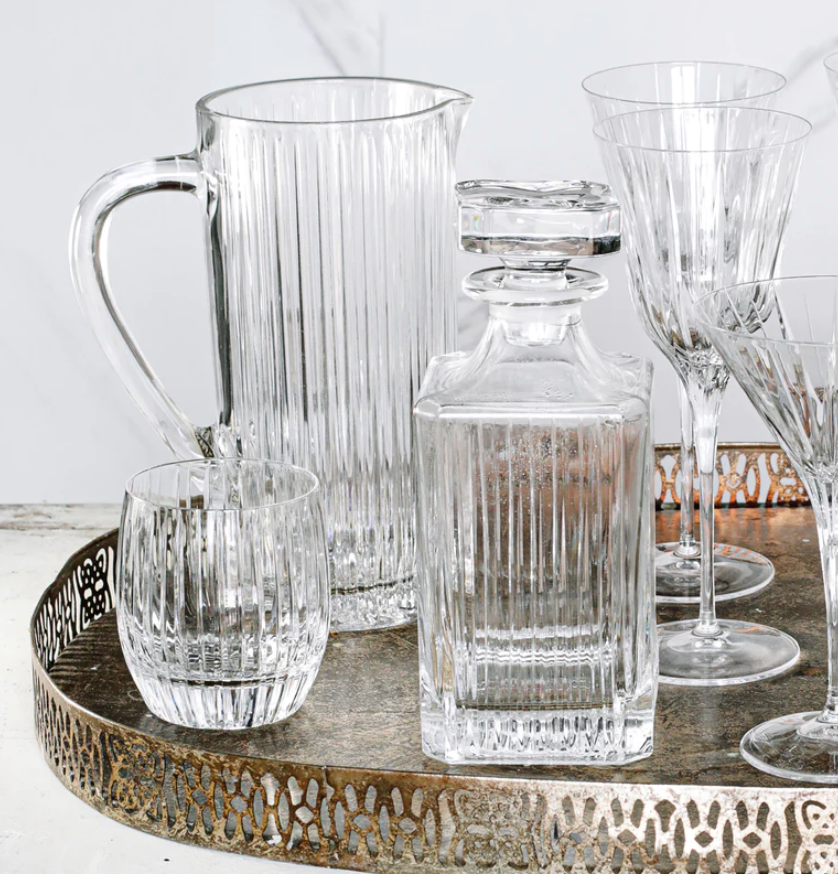 A metallic tray holds a Vietri Natalia Pitcher, a matching tumbler, a square decanter with stopper, and several tall stemmed glasses—all crafted in clear glass with elegant vertical ribbed patterns.