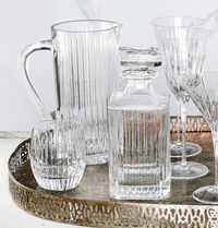 A metallic tray holds a Vietri Natalia Pitcher, a matching tumbler, a square decanter with stopper, and several tall stemmed glasses—all crafted in clear glass with elegant vertical ribbed patterns.