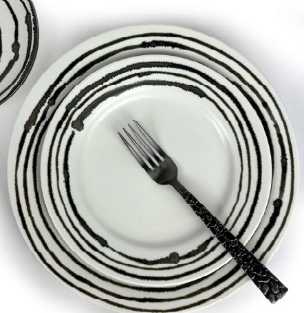 A black fork rests on a white Linea Dinnerware plate by Michael Wainwright, featuring concentric black rings. Another matching plate is partially visible in the upper left, all set against a white background.