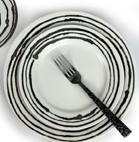 A black fork rests on a white Linea Dinnerware plate by Michael Wainwright, featuring concentric black rings. Another matching plate is partially visible in the upper left, all set against a white background.