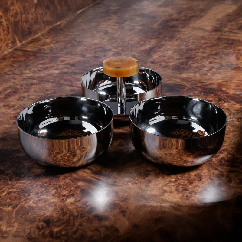 Three nickel bowls and a shaving brush with an amber handle from the Spector Condiment Set by Zodax are arranged on a brown marbled surface, the brush upright in the center bowl—a display echoing refined barware style.