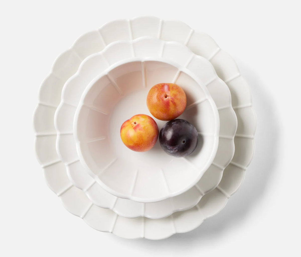 Three plums—two yellow-red and one dark purple—sit in a scalloped white bowl from the Iris Melamine Dinnerware Set of 4 by Blue Pheasant, stacked on two matching plates, all set against a white background.