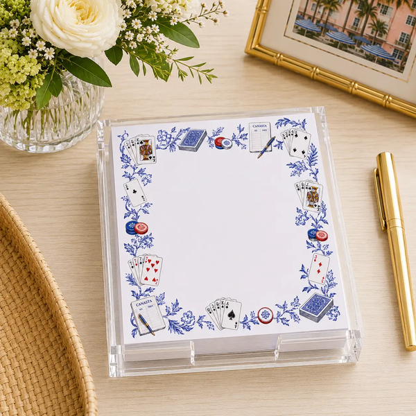 A clear acrylic holder containing a stack of paper for the Maven Home Proper Canasta Notepad & Holder, decorated with a blue floral border featuring illustrated playing cards, scorecards, and poker chips.