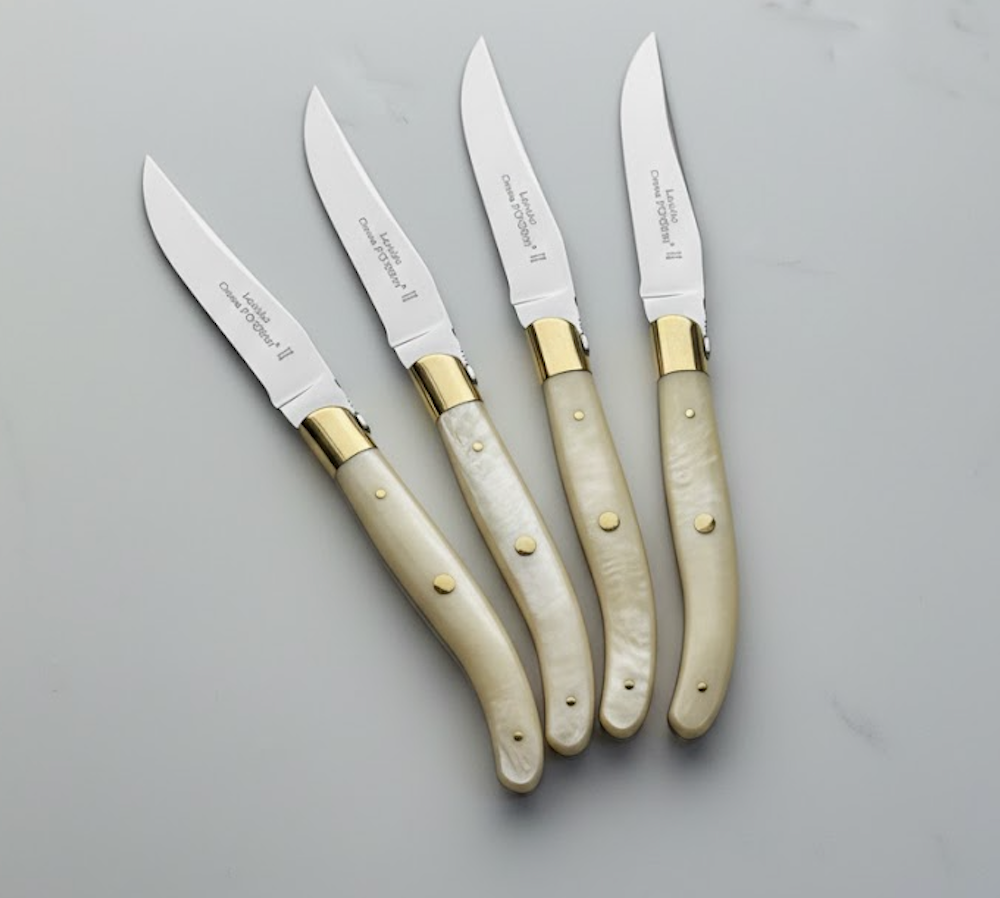 Four Claude Dozorme Laguiole Specialty Steak Knives, Brass Bolster Set/4 Ivory, are arranged diagonally on a light gray marble surface.