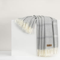 A light gray Lands Downunder Montauk Stripe Throw with a subtle plaid pattern and cream fringes is draped over a white surface. A small brown label accents one corner of this reversible Italian-made throw.