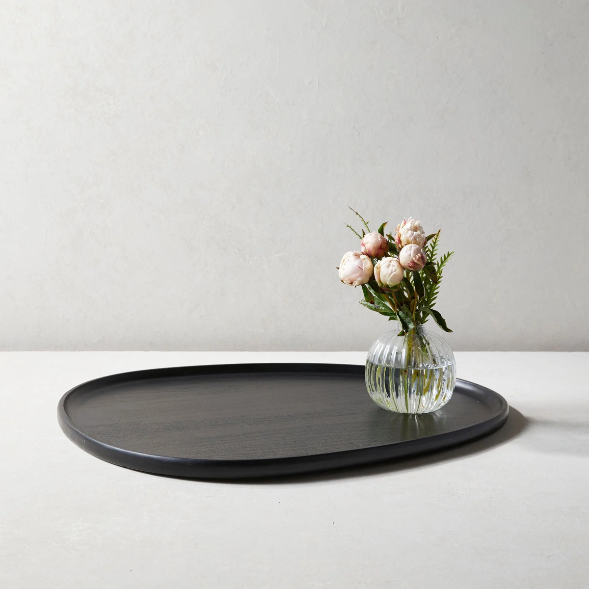 A round clear vase with pink peonies and greenery rests on the Be Home Tao Serving Tray Ebony XLarge, set against a plain light background.
