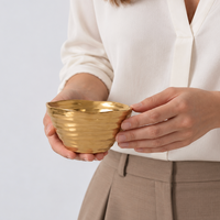 A person in a white blouse and beige pants holds the Dorrato Gold Bowl - by PAMPA BAY with both hands, showcasing its textured matte gold finish against a plain background.