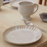 The Casafina Mallorca Salad Plate 9" in beige, featuring a scalloped edge and reactive glaze for a handcrafted, rustic look, sits on a light wooden table beside a matching mug, spoon, and gray napkin.
