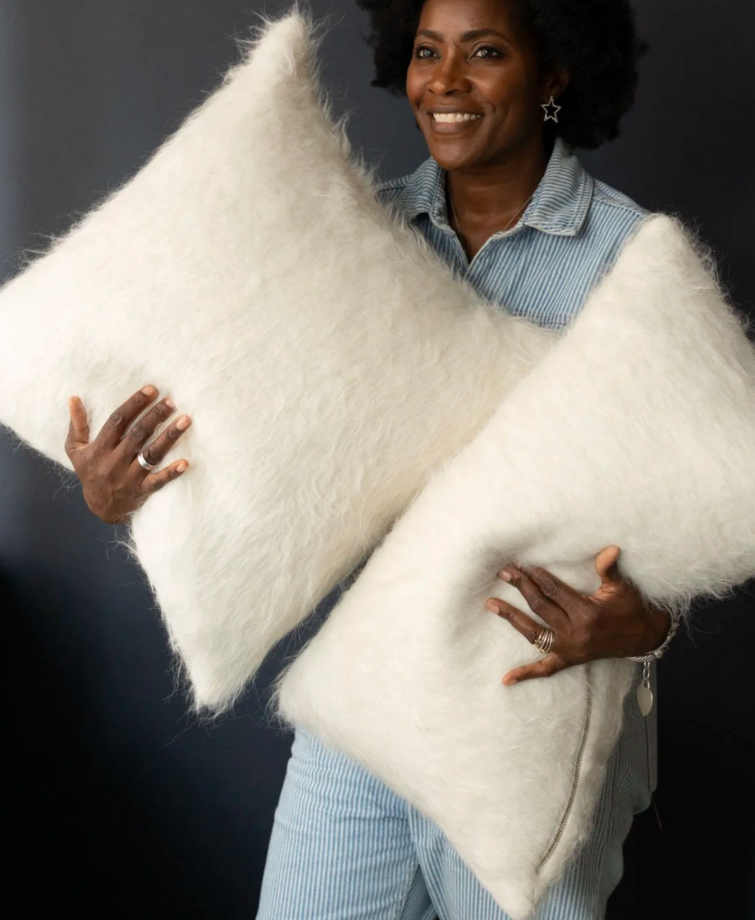A person in a light blue outfit smiles while holding two Dove & Donkey Woolly Mammoth Pillow Oat 20 x 20 pillows against a dark background.