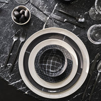 A modern, monochrome table setting featuring Serax’s Zuma Dinnerware Pacific—layered porcelain plates and a patterned bowl on black marble, paired with black cutlery, glasses, and black olives for a Bauhaus-inspired designer look.