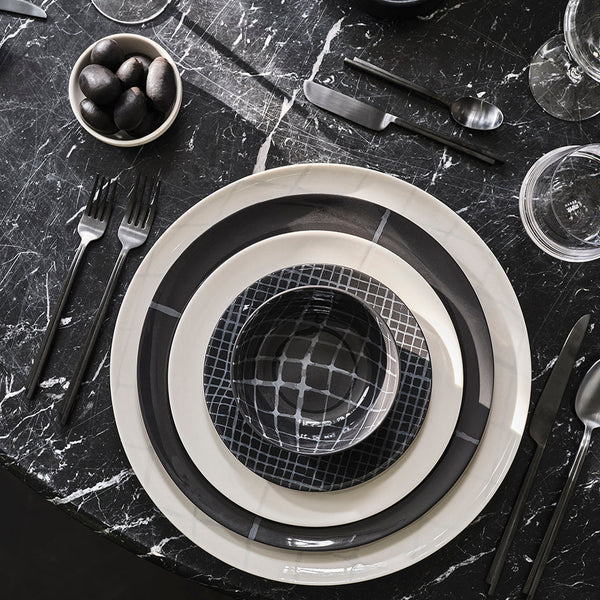 A modern, monochrome table setting featuring Serax’s Zuma Dinnerware Pacific—layered porcelain plates and a patterned bowl on black marble, paired with black cutlery, glasses, and black olives for a Bauhaus-inspired designer look.