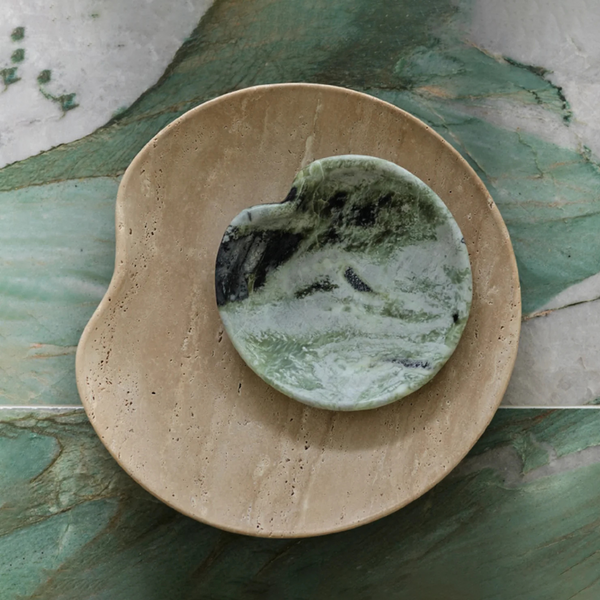 The Lily Marble Bowl Irish Green by Greg Natale sits atop a tan travertine dish, both arranged on a green and white marble surface—an elegant example of biomorphic design.
