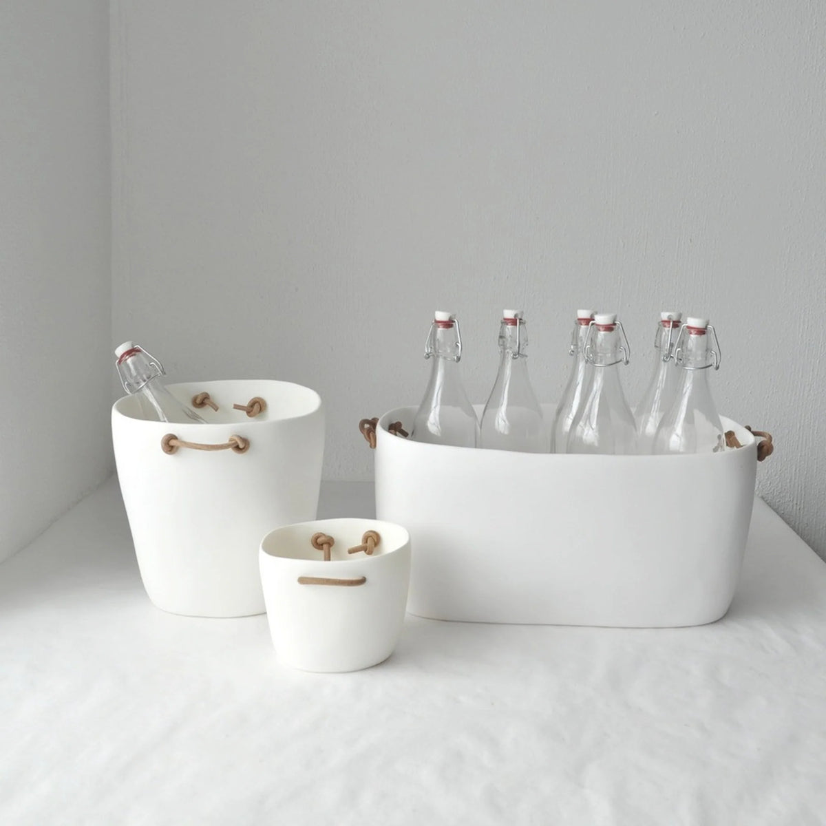 Three Tina Frey Champagne Buckets with leather handles are arranged on a white surface—two hold empty glass bottles with swing-top lids, and the smallest sits empty against a plain white wall.