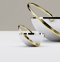Two reflective stainless steel Anna New York Ring Nut Bowls rest on a white surface, each surrounded by intersecting gold rings. Their modern, sculptural design makes them ideal for luxury home décor.