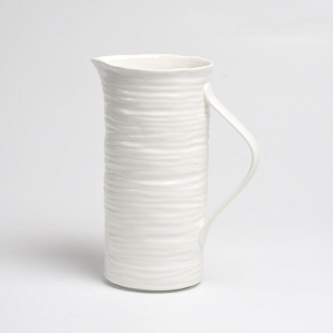 The Le Blanc Pitcher by PAMPA BAY, featuring a tall white ceramic build with a textured wavy surface and curved handle, makes an elegant centerpiece on a plain white background.