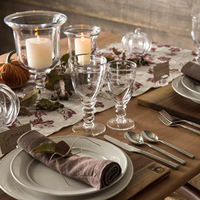 A rustic table features Simon Pearce Hartland Glass wine glasses, white plates on wood chargers, brown napkins with leaves, silver cutlery, candles in holders, and handwritten name cards on a patterned runner.