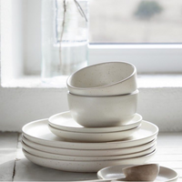 Casafina/Costa Nova Pacifica Salt Dinnerware—white stoneware plates and bowls with a matte finish—is stacked neatly on a countertop by a sunlit window, with a glass jar of water in the background.