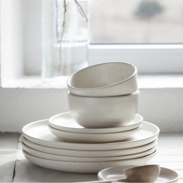 Casafina/Costa Nova Pacifica Salt Dinnerware—white stoneware plates and bowls with a matte finish—is stacked neatly on a countertop by a sunlit window, with a glass jar of water in the background.