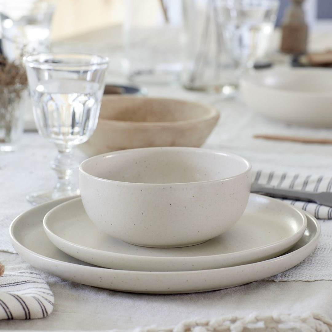 A close-up shows a Casafina/Costa Nova Pacifica Salt Dinnerware set—a bowl, salad plate, and dinner plate with a matte finish—stacked beside a clear water glass on a white tablecloth with striped napkins.
