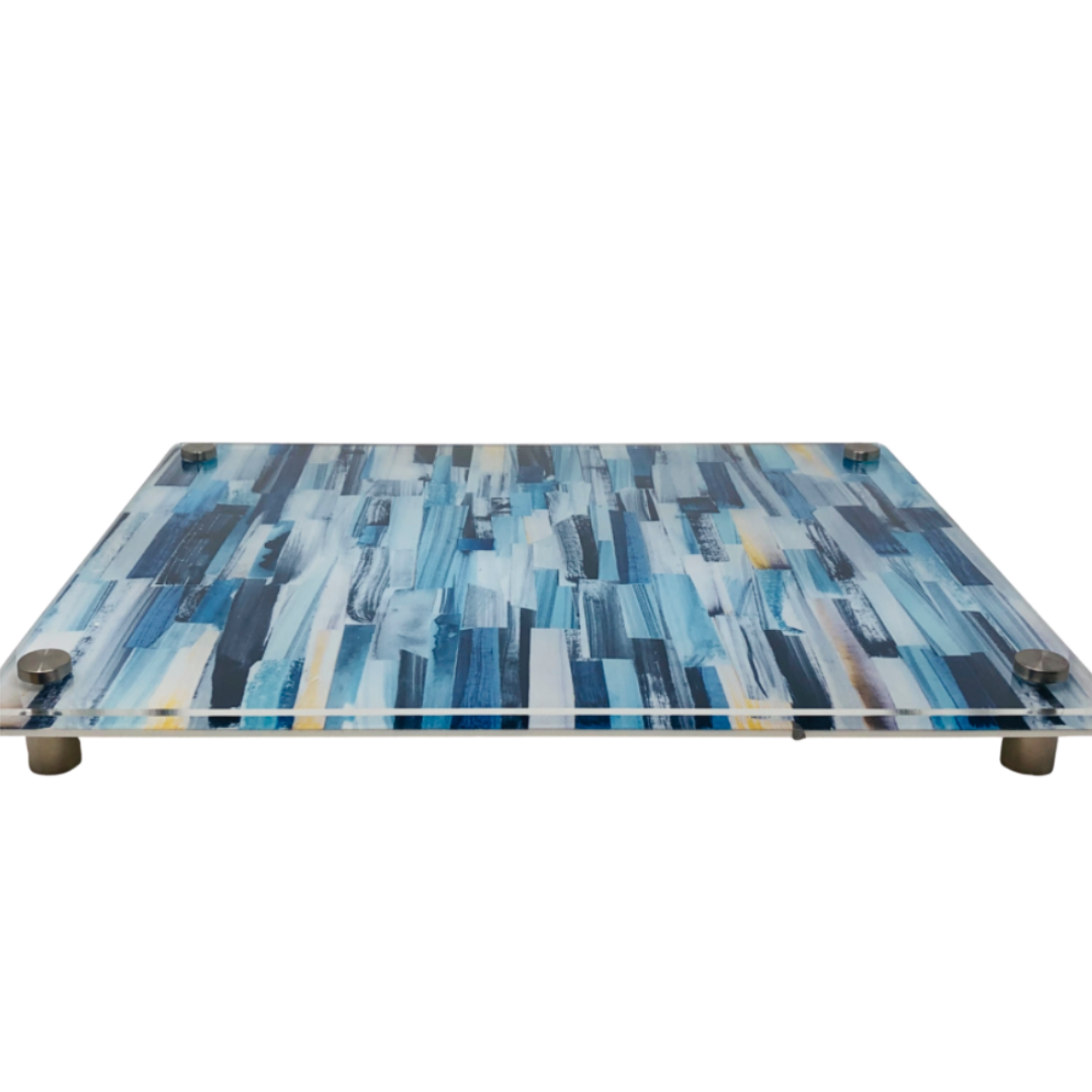 The Eye Candy Raised Serving Board Paint Stroke Blue features a rectangular blue design with white and yellow brushstroke accents, elevated by four metal corner feet.