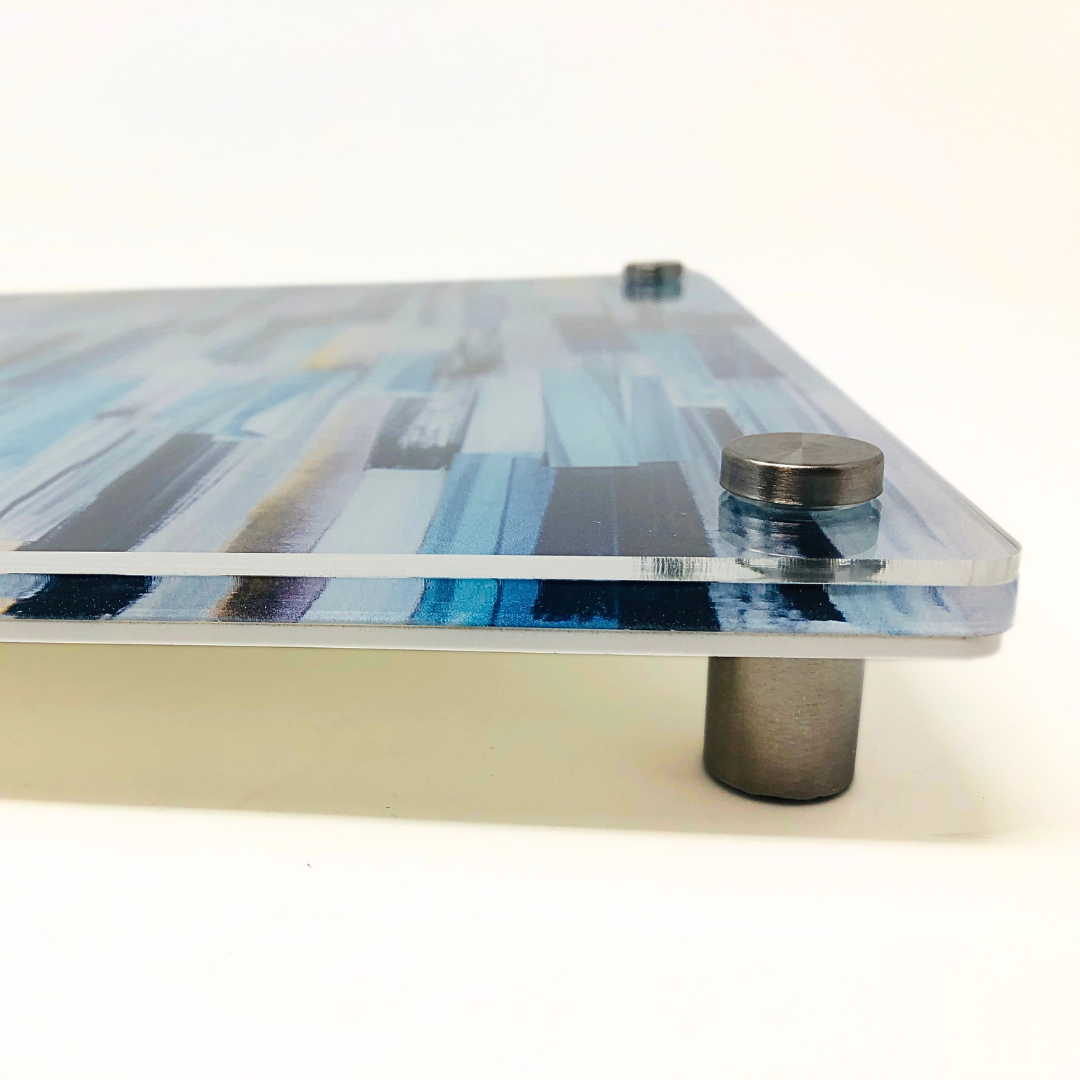 Close-up of the Eye Candy Raised Serving Board Paint Stroke Blue, a rectangular acrylic board featuring a blue abstract design, mounted on metal standoffs that accentuate its thickness and elevated position against a white background.